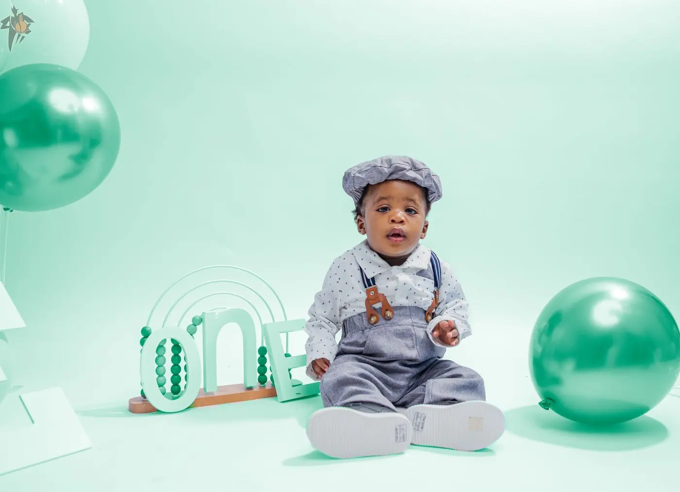 First birthday photography in Calgary: one-year-old boy in a flat cap and suspenders beside mint green ONE letters and balloons